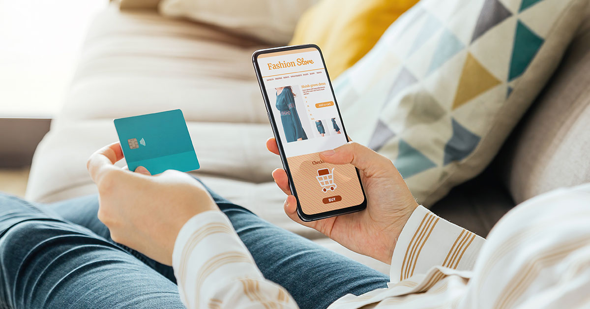 woman holding her credit card while shopping online using a mobile device. There is a blue dress on the screen.